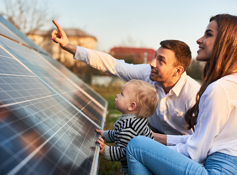 Man shows his family the solar panels on the plot near the house during a warm day. Young woman with a kid and a man in the sun rays look at the solar panels.